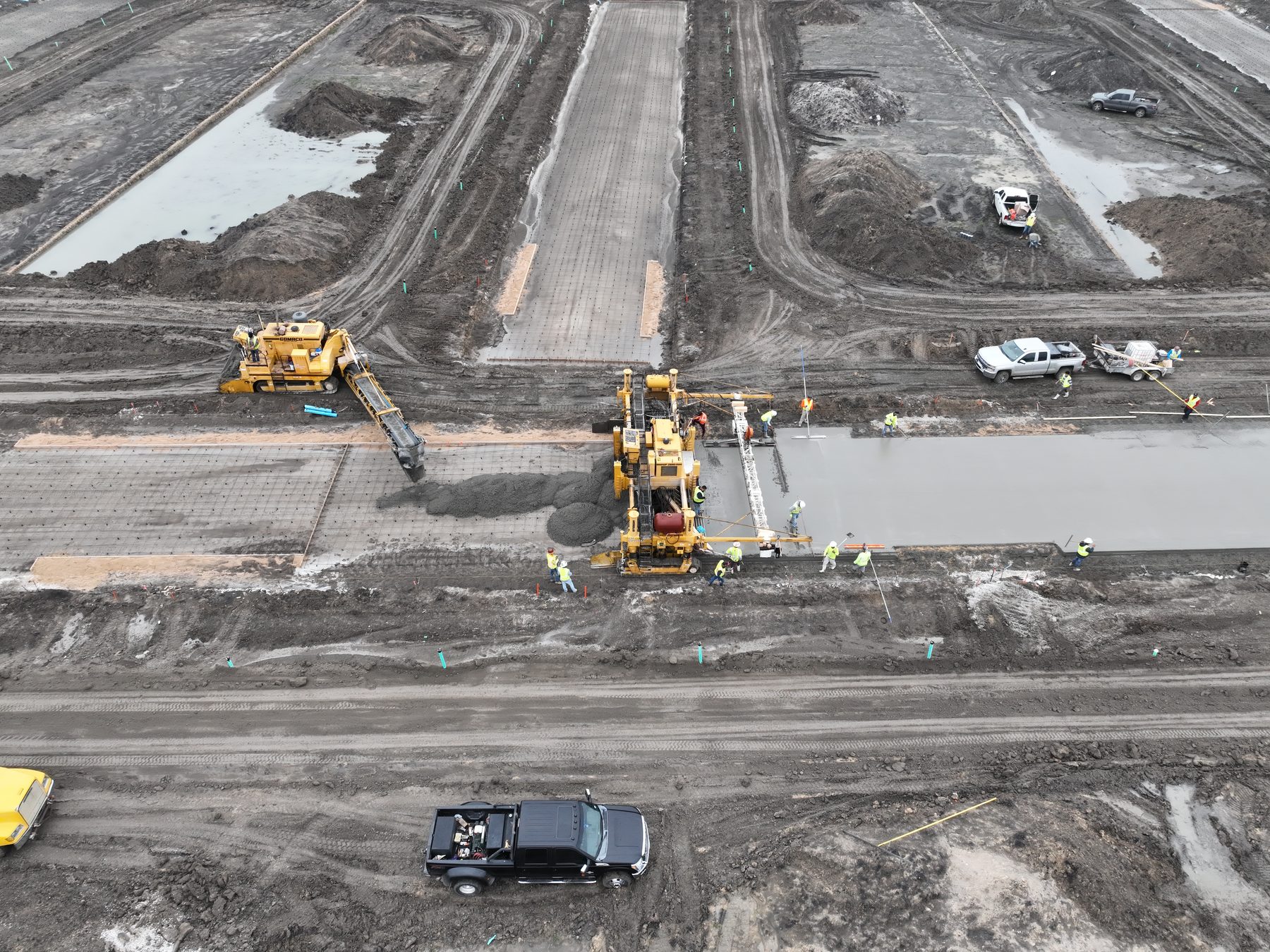 Aerial view of a paving train placing concrete at Arbor South in Princeton, Texas — slipform paver, concrete placer, and a crew in high-visibility gear working a fresh slab